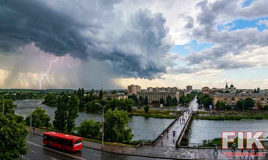 Панорамний вигляд Кропивницького під час наближення грози: темні штормові хмари та блискавка над річкою та містом, червоний автобус на дорозі, міст із пішоходами